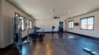 This fitness center is a thoughtfully designed, single-floor facility built to support a comprehensive workout experience. The space features polished dark concrete flooring complemented by interlocking rubber mat sections that define dedicated workout zones, providing both durability and comfort underfoot.

The cardio area is outfitted with multiple treadmills, stationary bikes, and a rowing machine, each positioned to take advantage of natural light streaming in through large, dark-trimmed windows. Wall-mounted flat-screen televisions are strategically placed throughout the room, allowing users to stay entertained during their workouts.

The strength training area includes a multi-use cable machine, a weight bench, a squat rack, and a dumbbell rack stocked with a range of free weights. A full-length wall mirror spans one side of the room, visually expanding the space and allowing for proper form checks during exercise.

Overhead, a large multi-blade ceiling fan serves as a central fixture, promoting air circulation throughout the facility. Recessed lighting provides consistent, even illumination across the entire space. A dedicated door leads out of the fitness area, and the open layout ensures ample room to move freely between equipment. The overall ambiance is clean, modern, and functional, making it a well-rounded facility for a variety of fitness routines.