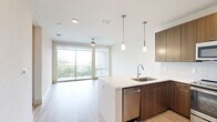 This well-appointed one-bedroom, one-bathroom unit offers a thoughtfully designed layout across a single floor. The open-concept kitchen and living area serves as the heart of the space, featuring rich walnut-toned cabinetry, a quartz waterfall peninsula with pendant lighting, stainless steel appliances including a range, microwave, refrigerator, and dishwasher, and a subtle tile backsplash. Light wood-style flooring runs throughout the main living area, enhancing the bright and airy atmosphere. Floor-to-ceiling sliding glass doors lead to a private balcony with scenic views of the surrounding landscape, while a ceiling fan adds comfort to the living room. The bedroom is carpeted and well-lit by a large window framing greenery and outdoor views, complemented by its own ceiling fan. A walk-in closet with dual hanging rods provides generous storage. The bathroom features large-format tile flooring, a soaking tub with tiled surround, a wood-toned vanity with a white countertop, and a backlit mirror. A hallway connects the private areas of the unit. Throughout, the design maintains a clean, contemporary aesthetic with a neutral palette and modern finishes.
