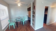 This single-floor space combines functionality and comfort in a well-organized layout. The dining area features a round glass-top table paired with vibrant teal metal chairs, adding a pop of color against the neutral walls and warm hardwood-style flooring that runs throughout the space. A ceiling fan with an integrated light fixture overhead provides both ventilation and ambient lighting.

Adjacent to the dining area is a compact kitchenette, equipped with dark cabinetry, a countertop workspace, and a sink. Upper dark cabinets provide storage, while the lower cabinetry maintains a cohesive, modern aesthetic. A small appliance, such as a coffee maker, is neatly placed on the counter, making the space practical for everyday use.

A built-in open shelving unit divides the kitchenette from the rest of the space, offering additional storage and display options. The shelves are white, creating a clean contrast with the darker cabinetry nearby. The overall ambiance is bright and tidy, with white walls and functional design elements that maximize the use of the available space efficiently.