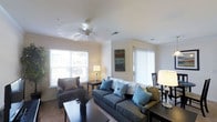 This single-floor residence is designed with a clean, functional layout that promotes an open and comfortable living environment. The kitchen is positioned along one wall, featuring a double-basin stainless steel sink, light-toned countertops, and white cabinetry offering ample storage. A half-wall divides the kitchen from the adjacent living area, creating an open-concept feel while maintaining a defined cooking space. A pendant light hangs above, adding a subtle decorative touch. Just off the kitchen, a doorway leads to a dedicated laundry room, providing added convenience with washer and dryer units visible within.

The living area is bright and inviting, benefiting from multiple large windows dressed with vertical blinds that allow generous natural light to fill the space. Crown molding lines the upper walls, adding a refined architectural detail. A ceiling fan with a light fixture is centrally mounted, contributing to both comfort and airflow. The room comfortably accommodates a seating arrangement and a dining area, making it well-suited for everyday living and entertaining. The overall aesthetic throughout is light, neutral, and well-maintained.