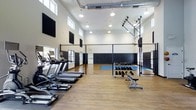 This fitness facility is designed to accommodate a wide range of athletic and wellness activities within a single, open-concept space. The interior is defined by dramatically high ceilings that create an airy, expansive atmosphere, complemented by rows of clerestory windows along the upper walls that allow generous natural light to fill the room throughout the day. Exposed industrial-style ductwork runs overhead, adding a modern utilitarian aesthetic to the bright white walls and warm hardwood-style flooring.

The facility is divided into distinct functional zones. A cardio area along one side houses multiple treadmills and elliptical machines, arranged to maximize usability. Opposite, a well-stocked dumbbell rack with an adjustable weight bench provides a dedicated free weights section. Black acoustic panels are strategically mounted on the walls, contributing to sound management during high-energy workouts.

A full chain-link-enclosed multi-sport court occupies the rear of the space, featuring a basketball hoop and a padded perimeter wall, making it suitable for court sports and dynamic training. The combination of thoughtful zoning, high-performance equipment, and architectural scale makes this a comprehensive and versatile fitness destination.