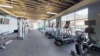 Located at 1225 Willowbrook Dr in Huntsville, Alabama, this fitness center offers a well-appointed, single-floor workout facility designed to support a comprehensive range of exercise routines. The space features an exposed ceiling with visible ductwork and structural beams, giving it an industrial aesthetic complemented by cool gray walls and durable rubber flooring throughout.

The cardio zone lines the perimeter wall of large windows, which flood the area with natural light and provide views of the outdoor pool and surrounding landscape. A row of Landice treadmills and elliptical machines are positioned to take full advantage of these views, enhancing the workout experience. Wall-mounted televisions are available for entertainment during exercise sessions.

The strength training area is well-stocked with SportsArt cable machines, a dumbbell rack, and dedicated resistance equipment, offering options for both isolated and compound movements. A functional training zone near the entrance features a power rack, a punching bag, kettlebells, and a BodyCraft rowing machine, catering to high-intensity and cross-training workouts. Stability balls and additional accessories are also available for flexibility and core training. Large glass entry doors provide a welcoming entrance and contribute additional natural light to the space.