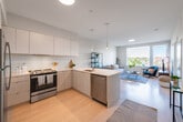 This thoughtfully designed single-floor residence showcases a clean, contemporary aesthetic throughout. The open-concept layout seamlessly connects the kitchen and living area, creating a bright, cohesive space ideal for both everyday living and entertaining. The kitchen is outfitted with stainless steel appliances — including a range, refrigerator, and dishwasher — complemented by flat-front cabinetry in a two-tone white and natural wood finish, a herringbone tile backsplash, and a white quartz peninsula with bar seating. Light hardwood flooring flows throughout the main living areas, enhancing the airy feel. The living room benefits from large windows that frame treetop and cityscape views, filling the space with abundant natural light. Two well-proportioned bedrooms each feature carpeted floors, neutral walls, and windows with peaceful outdoor vistas. The full bathroom includes large-format tile flooring, a walk-in shower, and a clean, functional layout. A dedicated storage area is also accessible from the hallway. Throughout the residence, a consistent palette of soft neutrals and natural materials creates a calm, modern atmosphere that is both practical and inviting.