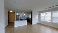 This contemporary single-floor space showcases a thoughtfully designed open-concept layout that seamlessly connects the kitchen and living areas. The kitchen is a standout feature, equipped with stainless steel appliances including a refrigerator, microwave, and dishwasher. Crisp white upper cabinetry contrasts elegantly with dark lower cabinets, while a granite-topped island with pendant lighting serves as both a functional workspace and a stylish focal point. A track lighting system overhead adds additional illumination throughout the kitchen area. Light hardwood flooring extends throughout the entire space, creating a warm and cohesive aesthetic. The open living area benefits from generous natural light streaming in through large windows fitted with blinds, offering views to an outdoor area. A glass door provides direct access to what appears to be a private balcony or patio, further enhancing the sense of openness and connectivity to the outdoors. The clean white walls and smooth ceilings contribute to a bright, airy ambiance throughout. The overall design reflects a modern sensibility with a neutral palette, making it a versatile and inviting space suitable for a variety of lifestyles.