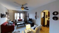 This single-floor home offers a welcoming and well-appointed interior defined by classic design details and a warm, comfortable ambiance. The living room serves as the heart of the space, furnished with plush sofas and an accent armchair arranged around a traditional dark-wood coffee table. Decorative touches, including wrought iron wall sconces and rich red accent pillows, lend the room a timeless, inviting character. A ceiling fan with a classic brass fixture provides both function and style overhead, while sheer curtain-draped windows allow soft natural light to filter into the room. A media console with a flat-screen television anchors one wall, offering a practical entertainment focal point. The living area transitions gracefully into an adjoining room through a distinctive arched doorway — an architectural detail that adds charm and visual interest to the layout. Warm hardwood flooring is visible in the connecting space, complementing the neutral tones of the main living area. The overall design balances comfort and classic style, making the space feel lived-in and welcoming throughout.