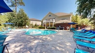Located at 412 Riverwood Dr in Crestview, Florida, this outdoor amenity area is centered around a generously sized freeform swimming pool with clear, sparkling blue water. The pool's curved design creates an inviting, resort-style atmosphere, complemented by a spacious concrete sun deck that wraps around the perimeter and is well-equipped with blue lounge chairs and patio umbrellas for shaded relaxation.

The adjoining clubhouse is a two-story structure featuring a classic combination of brick and siding exterior, topped with a pitched roof and a distinctive arched upper window. A covered upper-level balcony with white railings overlooks the pool area, while ground-floor glass doors provide direct access to the pool deck. The building is framed by well-maintained landscaping including trimmed hedges and mature trees.

On the opposite side of the pool, a lush canopy of tall trees provides natural shade and a tranquil, wooded backdrop, enhancing the sense of privacy and seclusion. The entire pool area is enclosed by a chain-link fence for safety. This well-appointed outdoor space offers a comfortable and visually appealing setting for leisure and recreation under the Florida sun.