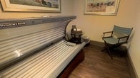 This single-level tanning space is designed to provide a comfortable and private experience for guests. The room is centered around a professional-grade tanning bed — identifiable as an Ergoline 245 model — equipped with a full array of UV lamps across both the upper canopy and lower bed surface, accommodating a full-body tanning session. The tanning bed's canopy is hinged open for easy access, showcasing its well-maintained interior. Beside the bed, a small round glass-top side table holds a wicker basket of tanning accessories and informational materials, keeping essentials conveniently within reach. The room features warm beige walls and rich wood-tone laminate flooring, contributing to a cozy, welcoming ambiance. Beach-themed framed artwork adorns the walls, depicting colorful beach huts and seaside scenes that reinforce a sunny, leisure-focused atmosphere. The overall design of the space is clean and functional, balancing the practical requirements of a professional tanning room with a relaxing aesthetic that helps guests feel at ease. The layout is efficiently organized to maximize comfort within the dedicated space.