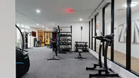 Located at 17-00 Pollitt Dr in Fair Lawn, NJ, this professionally equipped fitness center offers a comprehensive workout environment spread across a single, expansive floor. The space is thoughtfully divided into distinct functional zones, each designed to support a full range of exercise disciplines.

The main strength and cardio area features carpet flooring and is bathed in natural light through large, floor-to-ceiling windows that provide views of the surrounding outdoor environment. This zone is outfitted with a robust selection of True and Apex strength machines, adjustable benches, a multi-tiered dumbbell rack, kettlebells, and resistance training accessories. A wall-mounted flat-screen television adds convenience for users during workouts.

A dedicated cardio section houses multiple treadmills, a stair climber, and additional cardio machines, all arranged in an open, unobstructed layout beneath recessed ceiling lighting.

A separate studio area, enclosed by sleek black-framed glass partitions, features light hardwood flooring, floor-to-ceiling mirrors with warm wood framing, and a row of stationary cycling bikes. This studio provides an ideal environment for group fitness or individual cycling sessions, with a clean, modern aesthetic enhanced by pendant lighting and a full mirrored wall.

Throughout the facility, the design maintains a contemporary, minimalist character defined by white walls, clean lines, and an abundance of natural light, creating a bright and motivating atmosphere for all fitness levels.
