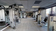 Located at 13 W Superior St in Chicago's vibrant River North neighborhood, this fitness center offers a well-appointed, single-floor workout facility designed to meet a wide range of training needs. The space is laid out in a long, open floor plan that promotes ease of movement between equipment zones, with light-toned carpet flooring and a drop tile ceiling fitted with recessed and panel lighting throughout.

The strength training area is extensively equipped with Life Fitness cable machines, weight benches, a dedicated free weights section featuring a full dumbbell rack, barbells, and weight plates, as well as a variety of selectorized machines for targeted muscle training. Full-length mirrors line portions of the walls, enhancing the sense of openness and allowing users to monitor their form during workouts.

A cardio section includes treadmills and stationary bikes, positioned to take advantage of large sliding glass doors and floor-to-ceiling windows that frame expansive views of the Chicago skyline. These windows flood the space with natural light and provide an energizing backdrop during workouts. Additional amenities include a workout bench, exercise mat storage, and private rooms accessible via dark wood-framed doors, likely serving as changing or utility spaces. The overall ambiance is clean, modern, and functional.