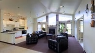 This single-story space is defined by its striking vaulted ceilings, which lend a sense of openness and grandeur to the interior. The main lounge area serves as the focal point of the space, centered around a classic fireplace framed by large clerestory windows that allow generous amounts of natural light to flood the room. The high-mounted windows provide bright, airy illumination while maintaining privacy.

The lounge is furnished with a cohesive set of tufted black leather sofas and armchairs arranged around a dark coffee table, creating a comfortable and welcoming seating arrangement. A large area rug anchors the seating group over the dark carpeted floor. Pendant light fixtures hang from the vaulted ceiling, supplementing the natural light. Decorative touches such as potted plants and floral arrangements add warmth to the space.

Adjacent to the lounge, a compact kitchen area is visible, equipped with standard appliances including a stove and overhead cabinetry, offering functional utility. Exit signage and a fire extinguisher indicate the space is designed with safety compliance in mind, suggesting a shared or community-use facility.