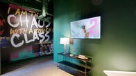 Located at 2801 Locust St in St. Louis, Missouri, this single-floor hallway space offers a generously proportioned lounge corridor that makes a lasting first impression through its distinctive, eclectic design. Deep forest-green walls set a dramatic backdrop throughout, complemented by polished concrete floors and exposed industrial ceilings with visible ductwork, conduit, and track lighting that reinforce a raw, urban aesthetic.

A central lounge area anchors the space, furnished with eye-catching metallic aviator-style egg chairs and a rich brown leather sofa, all arranged atop a bold geometric patterned area rug in warm orange, brown, and white tones. Built-in niche displays filled with stacked firewood flank a sleek wall-mounted electric fireplace, adding a rustic counterpoint to the industrial surroundings. A mounted flat-screen television above the fireplace enhances the lounge's functionality.

Large-scale, vibrant wall art featuring bold typographic graphics adds an energetic, expressive quality to the space. Structural columns, finished in matching deep green, frame the lounge area and contribute to the architectural rhythm of the corridor. Track and pendant lighting provides focused illumination throughout. The overall ambiance is dynamic and immersive—blending industrial, vintage, and artistic influences into a cohesive and memorable environment.