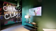 Located at 2801 Locust St in St. Louis, Missouri, this single-floor hallway space offers a generously proportioned lounge corridor that makes a lasting first impression through its distinctive, eclectic design. Deep forest-green walls set a dramatic backdrop throughout, complemented by polished concrete floors and exposed industrial ceilings with visible ductwork, conduit, and track lighting that reinforce a raw, urban aesthetic.

A central lounge area anchors the space, furnished with eye-catching metallic aviator-style egg chairs and a rich brown leather sofa, all arranged atop a bold geometric patterned area rug in warm orange, brown, and white tones. Built-in niche displays filled with stacked firewood flank a sleek wall-mounted electric fireplace, adding a rustic counterpoint to the industrial surroundings. A mounted flat-screen television above the fireplace enhances the lounge's functionality.

Large-scale, vibrant wall art featuring bold typographic graphics adds an energetic, expressive quality to the space. Structural columns, finished in matching deep green, frame the lounge area and contribute to the architectural rhythm of the corridor. Track and pendant lighting provides focused illumination throughout. The overall ambiance is dynamic and immersive—blending industrial, vintage, and artistic influences into a cohesive and memorable environment.