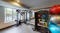 This fitness center is thoughtfully designed to support a wide range of workout routines within a single, open-plan floor. The space features durable speckled rubber flooring throughout, providing both traction and comfort during exercise. Recessed ceiling lighting ensures the room is well-lit, while large windows along one wall invite natural light and offer views of the surrounding outdoor area, creating an energizing atmosphere.

The cardio zone is equipped with multiple elliptical trainers, treadmills, a rowing machine, and a stationary bike, arranged to allow ample space between each unit. Wall-mounted flat-screen TVs are positioned for easy viewing during workouts. A dedicated strength training area features a TAG cable machine system, a full rack of dumbbells in graduated weights, and colorful medicine balls displayed on a tiered rack. Full-length mirrors line one wall, expanding the sense of space and allowing users to monitor their form.

Adjustable workout benches are available for a variety of strength exercises. The space also includes a sanitation station near the entrance for equipment hygiene. Framed artwork adds a finishing touch to the neutral-toned walls, one of which is accented in a deep teal-blue. The overall ambiance is clean, modern, and functional, making it a comprehensive facility for diverse fitness needs.