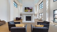 Located at 585 North Ave in New Rochelle, NY, this refined two-story communal space exemplifies upscale design with a seamless blend of classic architectural details and contemporary furnishings. The main level commands attention with its dramatic double-height ceilings, anchored by a grand white marble fireplace topped with a mounted flat-screen TV and flanked by built-in bookshelves. Multiple seating arrangements featuring tufted leather armchairs with accent pillows provide inviting gathering areas, while a formal dining setup occupies one corner of the space. Black-framed windows allow abundant natural light to pour in throughout both levels. A striking wrought-iron staircase with dark balusters connects the floors, serving as both a functional and decorative centerpiece. The upper level features an open mezzanine with decorative coffered ceilings accented in soft blue, additional lounge seating with outdoor views, and a glass-door passage to an adjoining space. Throughout both floors, neutral striped carpeting complements the crisp white wainscoting and walls. The overall ambiance is polished and welcoming, offering a versatile environment well-suited for both quiet relaxation and social gatherings.