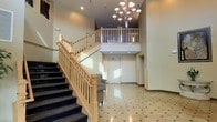 This impressive lobby makes a strong first impression with its grand, open design and refined architectural details. The centerpiece of the space is a sweeping staircase finished with light natural wood railings and dark-toned steps, rising to a mezzanine-level overlook that adds vertical drama and a sense of openness. Soaring double-height ceilings draw the eye upward to an elegant multi-arm chandelier, which serves as a striking focal point and bathes the space in warm, ambient light. The flooring is composed of polished tile in a classic geometric pattern, lending a timeless and sophisticated character to the entrance. Recessed lighting supplements the chandelier, ensuring the space is evenly and warmly illuminated throughout. A patterned upholstered accent chair is thoughtfully placed near the base of the staircase, offering a subtle residential touch within the otherwise formal setting. Double doors at the rear of the lobby provide access to additional areas beyond. The overall ambiance is welcoming yet distinguished, combining classic design elements such as the ornate chandelier and detailed railings with a clean, neutral color palette that keeps the space feeling bright, open, and professionally appointed.