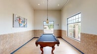 Located at 2710 Wilkie Dr in Rapid City, South Dakota, this billiards room is a well-appointed recreational space designed for leisure and social activity. The main area centers around a full-size pool table with a rich wood finish and classic claw-foot legs, set atop light hardwood flooring that runs throughout the space. Tall ceilings lend an airy, open feel, while a stylish tiered pendant light hangs directly above the table, providing focused illumination. The walls feature warm-toned tile wainscoting that extends along the lower portion, adding a polished, durable finish. Multiple large windows flood the room with natural light and offer views of an attractive outdoor landscape with rock features and mature greenery, enhancing the overall ambiance. Recessed lighting and wall art depicting billiard balls complete the inviting atmosphere. Adjacent to the main room is a fully equipped, ADA-accessible restroom finished with matching tile wainscoting, a granite-topped sink, large mirror, and grab bars, ensuring convenience and accessibility for all users. This thoughtfully designed space offers both functionality and comfort for recreation.