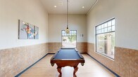 Located at 2710 Wilkie Dr in Rapid City, South Dakota, this billiards room is a well-appointed recreational space designed for leisure and social activity. The main area centers around a full-size pool table with a rich wood finish and classic claw-foot legs, set atop light hardwood flooring that runs throughout the space. Tall ceilings lend an airy, open feel, while a stylish tiered pendant light hangs directly above the table, providing focused illumination. The walls feature warm-toned tile wainscoting that extends along the lower portion, adding a polished, durable finish. Multiple large windows flood the room with natural light and offer views of an attractive outdoor landscape with rock features and mature greenery, enhancing the overall ambiance. Recessed lighting and wall art depicting billiard balls complete the inviting atmosphere. Adjacent to the main room is a fully equipped, ADA-accessible restroom finished with matching tile wainscoting, a granite-topped sink, large mirror, and grab bars, ensuring convenience and accessibility for all users. This thoughtfully designed space offers both functionality and comfort for recreation.