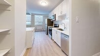 This well-appointed 3-bedroom, 1-bathroom unit offers a thoughtfully designed single-floor layout finished with light wood laminate flooring throughout, creating a clean and cohesive aesthetic. The open living room is generously proportioned, bathed in natural light from a wide bank of windows, and features a textured ceiling and a neutral color palette that lends a bright, airy feel to the space. The kitchen is a functional standout, outfitted with crisp white shaker-style cabinetry, a classic subway tile backsplash, and a full suite of stainless steel appliances including a refrigerator, electric range, built-in microwave, and dishwasher. A built-in shelving column divides the kitchen and living areas, adding practical storage while maintaining an open sightline. Each of the three bedrooms is comfortably sized, featuring large windows with outdoor views, individual radiator heating units, and light-toned walls. The primary bedroom includes a sliding-door closet for convenient storage. The bathroom is finished with glossy white subway tile surrounding a full soaking tub and shower, paired with a white vanity and undermount sink. Three hallways connect the rooms efficiently, contributing to the unit's well-organized and navigable floor plan.