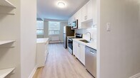 This well-appointed 3-bedroom, 1-bathroom unit offers a thoughtfully designed single-floor layout finished with light wood laminate flooring throughout, creating a clean and cohesive aesthetic. The open living room is generously proportioned, bathed in natural light from a wide bank of windows, and features a textured ceiling and a neutral color palette that lends a bright, airy feel to the space. The kitchen is a functional standout, outfitted with crisp white shaker-style cabinetry, a classic subway tile backsplash, and a full suite of stainless steel appliances including a refrigerator, electric range, built-in microwave, and dishwasher. A built-in shelving column divides the kitchen and living areas, adding practical storage while maintaining an open sightline. Each of the three bedrooms is comfortably sized, featuring large windows with outdoor views, individual radiator heating units, and light-toned walls. The primary bedroom includes a sliding-door closet for convenient storage. The bathroom is finished with glossy white subway tile surrounding a full soaking tub and shower, paired with a white vanity and undermount sink. Three hallways connect the rooms efficiently, contributing to the unit's well-organized and navigable floor plan.