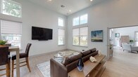 This well-appointed media room offers a thoughtfully designed communal space that seamlessly blends comfort and functionality. The room is defined by its impressively tall ceilings, which amplify the sense of openness and allow an abundance of natural light to pour in through the generously sized windows fitted with elegant roller shades. The main lounge area centers around a large wall-mounted flat-screen television paired with a sleek soundbar, with rich brown leather seating arranged over a soft area rug atop light wood-toned flooring throughout.

A striking accent wall clad in mixed-tone reclaimed wood planks serves as the backdrop for a built-in beverage bar, featuring an extended white quartz countertop, soft gray cabinetry, and an under-counter beverage refrigerator — ideal for hosting and casual gatherings. Adjacent to this, a casual dining nook with a wood-tone pub table and upholstered chairs sits beside bright windows overlooking a lush outdoor greenspace, offering a relaxed spot for conversation or a quick meal.

Curated artwork and tasteful decorative accents, including ceramic vases and sculptural pieces, add warmth and personality throughout the space. The overall aesthetic is clean, contemporary, and inviting, making this media room a versatile and sophisticated retreat.