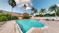 This single-story property is defined by its inviting outdoor amenities set within a lush, tropical environment. The centerpiece of the outdoor area is a generously sized swimming pool with clear turquoise water, framed by a well-maintained tiled pool deck equipped with lounge chairs and shaded umbrella seating — ideal for relaxing in the sun. The pool area is enclosed by a white fence, offering a sense of privacy while maintaining an open, airy feel. Towering palm trees and dense tropical foliage surround the space, creating a resort-like ambiance that evokes a warm, Florida-style setting. The exterior of the building features a warm coral-pink facade with teal shuttered windows, contributing to the property's distinctive tropical character. The overall outdoor environment is bright and cheerful, with abundant natural light filtering through the canopy of palms against an expansive sky. While interior images were not sufficiently illuminated to capture detail, the property's outdoor spaces make a strong impression, offering a comfortable and visually appealing setting enriched by mature landscaping and a well-appointed pool area suited for year-round enjoyment.