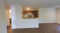This single-floor residence offers a practical and well-organized layout designed for comfortable everyday living. The open-concept design connects the living area to the kitchen, creating a sense of spaciousness and easy flow between the two spaces. Dark hardwood-style flooring runs throughout the living area, adding warmth and a classic aesthetic to the interior.

The kitchen is positioned behind a raised half-wall pass-through that doubles as a breakfast bar or serving counter, making it ideal for casual dining and entertaining. Rich, dark wood upper cabinetry provides ample storage and contributes to the kitchen's traditional character. A freestanding range with an overhead ventilation hood anchors the cooking area, while the white appliances and light countertops offer a clean contrast to the darker cabinetry.

The ceilings feature a textured finish, and flush-mounted lighting provides functional illumination throughout. A wall-mounted vent indicates a built-in HVAC system, ensuring year-round climate comfort. The white walls throughout the space create a bright, neutral backdrop that allows for personalization and versatile décor choices. Overall, this residence combines functional design with a warm, inviting ambiance suited for a variety of lifestyles.