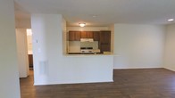 This single-floor residence offers a practical and well-organized layout designed for comfortable everyday living. The open-concept design connects the living area to the kitchen, creating a sense of spaciousness and easy flow between the two spaces. Dark hardwood-style flooring runs throughout the living area, adding warmth and a classic aesthetic to the interior.

The kitchen is positioned behind a raised half-wall pass-through that doubles as a breakfast bar or serving counter, making it ideal for casual dining and entertaining. Rich, dark wood upper cabinetry provides ample storage and contributes to the kitchen's traditional character. A freestanding range with an overhead ventilation hood anchors the cooking area, while the white appliances and light countertops offer a clean contrast to the darker cabinetry.

The ceilings feature a textured finish, and flush-mounted lighting provides functional illumination throughout. A wall-mounted vent indicates a built-in HVAC system, ensuring year-round climate comfort. The white walls throughout the space create a bright, neutral backdrop that allows for personalization and versatile décor choices. Overall, this residence combines functional design with a warm, inviting ambiance suited for a variety of lifestyles.