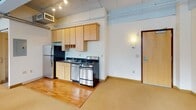 Located at 814 Hennepin Ave in the heart of downtown Minneapolis, this urban loft-style space blends industrial character with functional design across a single floor. The main living area is defined by soaring exposed concrete ceilings with visible ductwork and beam details, lending an authentic industrial aesthetic. A row of tall windows along multiple walls floods the space with natural light while framing views of the surrounding downtown Minneapolis skyline and streetscape. Flooring transitions between warm hardwood and carpet, adding texture and delineating different areas within the open layout. The kitchen is compact yet functional, featuring light wood cabinetry, a stainless steel range, refrigerator, and a dark-tiled backsplash. Track lighting illuminates the kitchen area, complementing the natural light throughout. The bathroom is clean and straightforward, offering a full soaking tub and shower combination, a pedestal sink, and classic round mirror. Exposed HVAC elements throughout the space reinforce the industrial loft aesthetic. The open floor plan allows for flexible use of the space, with ample room for varied configurations. This address places the space within walking distance of Minneapolis's vibrant arts, dining, and entertainment district along Hennepin Avenue.