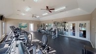 This fitness center is a thoughtfully designed, single-floor facility built to support a full range of workout routines. The space features soaring vaulted ceilings that lend an open, airy feel, while large windows along the perimeter flood the room with natural light and offer views of an outdoor pool area. Ceiling fans are mounted throughout to maintain comfortable airflow during workouts.

The flooring consists of durable dark rubber matting, ideal for both cardio and strength training use. The cardio zone is well-stocked with TRUE-brand treadmills, elliptical trainers, and recumbent bikes, each equipped with individual consoles. Multiple wall-mounted flat-screen televisions are positioned throughout for entertainment during workouts.

The strength training area includes a cable machine, adjustable weight benches, a weight rack with free weights, and a medicine ball station. Full-length mirrors are strategically placed along the walls to allow for form monitoring and to further enhance the sense of space.

The décor is clean and modern, with neutral warm-toned walls accented by colorful abstract artwork. The facility is accessed through glass-paned doors and includes clearly marked emergency exits, ensuring both convenience and safety. Overall, the space offers a professional and motivating environment for fitness enthusiasts.