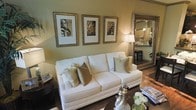 This single-floor space exudes a refined, welcoming atmosphere anchored by a thoughtfully designed living area. The room centers around a plush white sofa dressed with sage green and olive accent pillows, creating a soft, neutral palette that feels both sophisticated and livable. Warm golden-toned walls enhance the cozy ambiance, complemented by rich dark hardwood flooring that adds depth and contrast throughout.

A large ornate gold-framed mirror is mounted prominently on one wall, visually expanding the space while reflecting light from a classic table lamp with a warm glow. Three matching gold-framed sepia-toned architectural prints are displayed above the sofa, adding a curated, gallery-like quality to the room.

Decorative accessories on the coffee table, including a floral arrangement and elegant figurines, contribute to the polished aesthetic. A glimpse through the mirror reveals an adjacent kitchen area, suggesting a connected, open layout. The overall design balances classic elegance with comfortable livability, making it an inviting and well-composed living environment.
