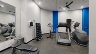 Located at 309 Oak St SE in Minneapolis, MN, this fitness center is a single-floor exercise room designed to deliver a comprehensive workout experience within a compact footprint. The space features a clean, modern aesthetic with white walls accented by a bold blue vertical stripe, a drop ceiling with recessed lighting and ceiling fans for ventilation, and durable speckled rubber flooring suited for high-traffic athletic use.

The room is thoughtfully equipped with a variety of cardio and strength training options. Cardio equipment includes a Cybex treadmill, a stationary spin bike, and an elliptical machine. For strength training, the space offers a rack of dumbbells spanning multiple weight increments, a multi-function cable and pulley machine, and a dedicated storage rack holding an assortment of medicine balls and stability balls. A weight bench provides additional versatility for various exercises.

Two wall-mounted flat-screen televisions are positioned near the entrance, offering entertainment during workouts. The entry area also features a glass door with an informational panel and additional step platforms stored nearby. Mirrors along one wall help maximize the sense of space and allow users to monitor their form. Overall, this fitness center offers a well-rounded selection of equipment in an efficiently organized, clean, and functional environment.