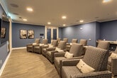 This single-floor media and screening room is thoughtfully designed to deliver a comfortable, communal viewing experience. The space is defined by its soothing blue-painted walls, crisp white crown molding, and light wood-look flooring, which together create a welcoming and polished atmosphere.

Rows of plush, patterned recliner-style armchairs are arranged in a gentle arc facing the focal wall, which features a large wall-mounted flat-screen television flanked by retractable roller shades — ideal for light control during screenings. The chairs are upholstered in a neutral herringbone-style fabric and accented with white throw pillows, offering both comfort and a cohesive aesthetic. Small side tables are positioned throughout for added convenience.

Recessed ceiling lighting provides even, ambient illumination throughout the room, complemented by a subtle blue accent light along the front wall that adds a cinematic touch. In-ceiling speakers suggest integrated audio capabilities for an immersive experience.

Decorative botanical grid artwork adorns one wall, while framed black-and-white photography adds visual interest near the entrance. A small refreshment station is tucked into the corner, completing the room's amenities. A glass-paneled door provides access to an adjacent space, enhancing the room's connectivity and functionality.