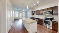Located at 500 Chris Kelley Blvd in Hutto, TX, this well-appointed one-bedroom, one-bathroom unit offers a thoughtfully designed layout across a single floor. The open-concept kitchen and living area serves as the heart of the space, showcasing rich dark-wood cabinetry, a striking granite-topped center island with pendant lighting overhead, and a full suite of stainless steel appliances including a refrigerator, range, microwave, and dishwasher. A tile backsplash and recessed lighting add polished finishing touches, while warm hardwood-style flooring extends throughout the main living area, enhancing the sense of openness. Large windows at the far end of the living space invite natural light and provide outdoor views, complemented by a ceiling fan for added comfort. The bedroom offers plush carpeting, a ceiling fan, and a generously sized window that frames pleasant outdoor scenery, creating a calm and restful retreat. The bathroom is finished with neutral tile flooring, a granite vanity with dark wood cabinetry, a framed mirror, and a full tub-shower combination with tile surround. Clean lines, neutral tones, and consistent finishes throughout give the space a cohesive and modern aesthetic.