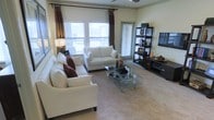 This single-floor space offers a thoughtfully designed living area that combines comfort and modern style. The living room is a standout feature, bathed in natural light streaming through multiple large windows dressed with elegant drapes and adjustable blinds. The neutral, warm tones of the walls and plush carpeted flooring create a welcoming ambiance throughout.

The living area is generously proportioned, accommodating a full seating arrangement that includes a large sofa and a loveseat, both upholstered in crisp white fabric and accented with rich burgundy throw pillows. A sleek glass-top coffee table anchors the seating area, adding a touch of contemporary flair. On one wall, a dark-toned media console and an open display shelving unit provide ample storage and display space, complementing the room's overall aesthetic.

A ceiling fan is centrally positioned overhead, contributing to both the comfort and functionality of the space. The room also features access to what appears to be a private outdoor area through a glass door, further enhancing the sense of openness. The cohesive design, characterized by its blend of neutral tones, dark wood accents, and carefully selected furnishings, gives the space a polished, move-in-ready appeal.