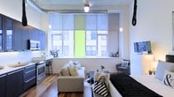 This single-floor space showcases an open-concept layout that integrates the living area and kitchen into one cohesive environment. The design draws on industrial influences, evident in the exposed ductwork along the ceiling and the warm hardwood flooring that runs throughout. A large bank of windows along one wall floods the interior with abundant natural light, offering views of the surrounding urban streetscape. Roller shades in neutral tones, accented by a bold lime green panel, provide flexible light control while adding a pop of color to the space. The living area is furnished with a plush sofa, coordinating accent chair, and a central coffee table, creating a comfortable and stylish gathering zone. A cowhide rug and contemporary accessories contribute to the eclectic, modern aesthetic. The kitchen area is visible in the background, featuring dark cabinetry, a stainless steel appliance suite, and a raised bar counter with seating, making it functional for both cooking and casual dining. A ceiling fan centered above the main living space enhances air circulation. Overall, the space delivers a well-balanced blend of industrial character and contemporary comfort.