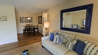 This single-floor residence showcases an open-concept design that seamlessly connects the living and dining areas into one cohesive, functional space. The living area is anchored by a neutral-toned sofa accented with blue and grey patterned throw pillows, complemented by a large ornate dark-framed mirror mounted on the wall that adds depth and visual interest to the room. A glass-topped coffee table and table lamp with a warm glow contribute to the inviting ambiance.

Rich hardwood flooring extends throughout the space, providing a warm and consistent foundation that unifies the living and dining zones. The dining area is furnished with a round dark wood table and matching chairs, topped by a pendant light fixture that draws the eye upward and defines the space. A gallery-style wall art arrangement adds a personalized, decorative touch to the dining area backdrop.

Neutral beige walls provide a versatile canvas that allows the blue accent pieces and dark wood furnishings to stand out. The layout offers an efficient flow between zones, making the space feel both comfortable and well-organized for everyday living and entertaining.