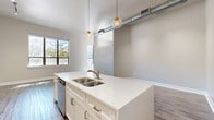 This single-floor unit offers a thoughtfully designed open-concept layout that seamlessly connects the kitchen and living area. The space features rich, dark hardwood-style flooring throughout, complemented by neutral warm-gray walls and crisp white trim that create a clean, modern aesthetic. Soaring ceilings with exposed industrial ductwork add a distinctive urban character, while globe pendant lights over the kitchen island provide warm, stylish illumination alongside a track lighting system.

The kitchen is a standout feature, centered around a large island with a white quartz countertop and a double stainless steel sink. White shaker-style cabinetry extends to the ceiling, offering generous storage, and is paired with a full suite of stainless steel appliances including a refrigerator with ice maker, electric range, microwave, and dishwasher. The open living area benefits from multiple windows that invite natural light and offer views of the surrounding neighborhood.

The two bathrooms are finished with white cabinetry, quartz countertops, brushed nickel fixtures, and tile-surround walk-in showers. A dedicated laundry room accommodates a full-size washer and dryer with additional shelving. The closet provides double hanging rods for organized storage, and a hallway connects all areas of the unit efficiently.