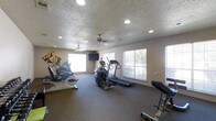 This single-floor fitness room offers a dedicated space for exercise and physical wellness. The room is outfitted with a selection of cardio equipment, including a treadmill, elliptical trainer, and stationary bike, providing users with a variety of workout options. A weight bench with free weights is also available for strength training. The flooring consists of durable, dark carpet well-suited for a fitness environment.

The space is illuminated by a combination of recessed ceiling lights and natural light streaming in through multiple windows along the walls, creating a bright and energizing atmosphere. Two ceiling fans are mounted overhead, promoting air circulation throughout the workout area. A wall-mounted flat-screen television adds a layer of entertainment for users during exercise sessions.

The walls are painted in a neutral tone, complementing the textured ceiling. A decorative piece of wall art featuring a colorful musical motif adds a subtle visual accent to the room. Overall, the space is thoughtfully arranged to maximize usability while maintaining an open, uncluttered feel conducive to an effective workout experience.