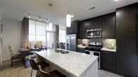 This single-floor space showcases a contemporary open-concept layout that seamlessly integrates the kitchen and living areas. The kitchen is a standout feature, anchored by a generous island topped with striking white and grey veined stone countertops that provide both prep space and casual seating for bar-height chairs. A stainless steel undermount sink sits centrally within the island, complemented by cylindrical pendant lights that hang above, adding a refined decorative touch.

Dark espresso cabinetry lines the kitchen walls, extending to the ceiling to maximize storage. These contrast sharply with the light countertops and a neutral tile backsplash. Stainless steel appliances, including a French door refrigerator, gas range, and built-in microwave, complete the culinary space with a polished, modern aesthetic.

The open living and dining area beyond the kitchen benefits from floor-to-ceiling windows that flood the space with natural light and offer expansive outdoor views. Light wood-toned flooring runs throughout, tying the spaces together cohesively. Track lighting adds layered illumination, enhancing the overall bright and airy ambiance of this well-appointed, modern home.