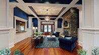Located at 1 Legacy Oaks Place in Asheville, NC, this single-floor space exudes refined Southern charm through a seamless blend of traditional craftsmanship and upscale design. The grand entryway welcomes visitors through stately wood-and-glass double doors framed by transom windows, flooding the interior with natural light. Rich, warm hardwood flooring extends throughout the open layout, anchoring the space with a sense of warmth and continuity. Striking navy blue walls are balanced by crisp white wainscoting, decorative pilasters, and beadboard paneling that line the lower walls, creating a classic, sophisticated aesthetic. The coffered ceiling, accented with recessed lighting and a statement orb chandelier, adds architectural depth and grandeur. A dramatic floor-to-ceiling stacked stone fireplace serves as the room's focal point, complete with a rustic wood mantel and a raised stone hearth. A service window integrated into one wall suggests a reception or hospitality function, adding practical elegance. The space is thoughtfully arranged with multiple seating areas, conveying both comfort and versatility. Overall, this beautifully appointed space offers a polished, welcoming atmosphere ideal for gatherings and community engagement.