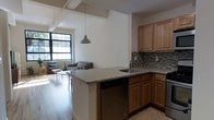 This single-floor space showcases a thoughtfully designed open-concept layout that seamlessly connects the kitchen and living areas. The kitchen is a standout feature, offering warm honey-toned wood cabinetry, granite countertops, and a mosaic tile backsplash that adds texture and visual interest. A peninsula with an integrated sink and dishwasher provides both additional workspace and a natural divider between the kitchen and living areas, while a pendant light overhead adds a refined focal point. The hardwood flooring flows throughout, lending warmth and continuity to the space. Exposed structural beams on the ceiling contribute an industrial character, complementing the clean, modern aesthetic. Large windows in the adjacent living area allow generous natural light to fill the space, creating a bright and welcoming ambiance. The living area offers a comfortable, open setting well-suited for relaxing or entertaining. The combination of modern finishes, open sightlines, and abundant natural light makes this a versatile and inviting space that balances both function and style.
