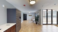 This two-floor residential building greets residents and visitors with a well-appointed lobby designed for both function and comfort. The entrance area is finished with large-format ceramic tile flooring in warm earth tones, providing a clean and durable surface throughout the common space. Flush-mount ceiling fixtures provide consistent ambient lighting, complementing the natural light that floods in through expansive windows along the exterior wall.

A prominent bank of modern, dark-toned package and mail lockers lines one side of the lobby, offering secure and convenient parcel retrieval for residents. A dedicated seating nook features a cushioned armchair arranged on a soft area rug, accompanied by a decorative potted plant and framed wall art, creating an inviting space to pause or wait. The neutral gray wall tones and white trim lend the lobby a clean, contemporary aesthetic.

A secured interior door provides controlled access to the rest of the building, ensuring resident safety and privacy. The textured ceiling adds a subtle architectural detail consistent throughout the common area. Overall, the lobby strikes a balance between practicality and welcoming design, setting a polished and organized tone for the two-story building it serves.