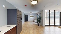 This two-floor residential building greets residents and visitors with a well-appointed lobby designed for both function and comfort. The entrance area is finished with large-format ceramic tile flooring in warm earth tones, providing a clean and durable surface throughout the common space. Flush-mount ceiling fixtures provide consistent ambient lighting, complementing the natural light that floods in through expansive windows along the exterior wall.

A prominent bank of modern, dark-toned package and mail lockers lines one side of the lobby, offering secure and convenient parcel retrieval for residents. A dedicated seating nook features a cushioned armchair arranged on a soft area rug, accompanied by a decorative potted plant and framed wall art, creating an inviting space to pause or wait. The neutral gray wall tones and white trim lend the lobby a clean, contemporary aesthetic.

A secured interior door provides controlled access to the rest of the building, ensuring resident safety and privacy. The textured ceiling adds a subtle architectural detail consistent throughout the common area. Overall, the lobby strikes a balance between practicality and welcoming design, setting a polished and organized tone for the two-story building it serves.