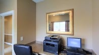 This single-floor business center offers a practical and well-appointed workspace designed to meet essential professional needs. The room is finished in warm, neutral earth tones that create a calm and focused atmosphere. A granite-topped workstation serves as the centerpiece, equipped with a desktop computer and a multifunction printer, providing guests or occupants with convenient access to computing and printing capabilities. A large, ornately framed gold mirror is mounted on the wall above the desk, visually expanding the space and reflecting natural light that filters in through a window dressed with rich, dark curtain panels. The overall design balances functionality with understated elegance, incorporating details such as a leather-upholstered chair and clean, uncluttered wall surfaces. The layout is efficient, making thoughtful use of the available space to ensure all essential business tools are within easy reach. The combination of practical furnishings and decorative accents gives the room a polished, professional character suited for productivity.