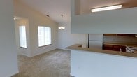 This single-floor space showcases an open-concept design that seamlessly connects the kitchen and living areas. The vaulted ceiling adds a sense of spaciousness and volume to the main living area, which is finished with neutral-toned wall-to-wall carpeting that contributes to a warm, comfortable atmosphere. A decorative chandelier hangs as a focal point over the living space, complementing the airy feel created by the elevated ceiling line.

The kitchen features a wraparound peninsula with a light-toned countertop, offering both additional workspace and a natural visual divider between the cooking and living zones. A full-size refrigerator is visible within the kitchen area, and the cabinetry provides ample storage. The kitchen's upper structure extends toward the ceiling, creating an architectural detail that adds depth to the space.

Natural light filters into the living area through a window fitted with horizontal blinds, brightening the neutral color palette of white walls and beige carpet. Overhead lighting in the kitchen ensures the workspace remains well-lit. Overall, the space presents a clean, functional, and open environment suited for everyday living.