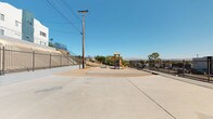 This outdoor recreational space is an open-air playground area set within a desert-climate environment, fully enclosed by iron perimeter fencing that provides a secure boundary. The space is divided between a wide, flat concrete surface suitable for open play and a sand-based playground zone at its center. The playground features multiple freestanding play structures, including two tower units with covered rooftops, yellow and blue slides, climbing bars, and a horizontal ladder bridge with connecting rails — offering varied physical activities for children. The structures are constructed with metal framing in blue and purple tones, accompanied by rubber-matted ground panels beneath key areas for safety. Surrounding the playground, sparse desert-adapted shrubs and small trees add minimal greenery to the otherwise arid landscape. A utility pole stands within the enclosure, and the surrounding neighborhood includes multi-story residential buildings visible beyond the fencing. The backdrop features distant mountain views beneath a clear blue sky, contributing to the open, sun-exposed character of the space. The overall setting reflects a functional, utilitarian design intended to serve a residential community.