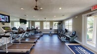 Located at 5525 Elkhorn Dr in Indianapolis, IN, this well-appointed fitness center is a single-floor facility designed to support a comprehensive workout experience. The space is generously sized, with a thoughtful layout that separates cardio and strength training zones for ease of use.

The cardio area is lined with a row of treadmills and elliptical trainers, complemented by stationary and recumbent bikes. Large windows along multiple walls flood the space with natural light, while ceiling fans and recessed lighting maintain a comfortable, well-lit environment. Wall-mounted flat-screen televisions are positioned for viewing during cardio sessions.

The strength and functional training area features a multi-station cable machine, a full rack of dumbbells, a weight bench, battle ropes, a punching bag, kettlebells, stability balls, and foam rollers — offering a diverse range of training options. Durable rubber flooring throughout provides safety and support for all exercise types.

French doors on one side of the facility open to the outdoors, enhancing ventilation and providing a seamless connection to the surrounding grounds. The textured ceiling, neutral gray walls, and clean white trim give the space a polished, welcoming aesthetic. This fitness center is well-suited to serve as an amenity for a residential community.
