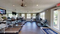 Located at 5525 Elkhorn Dr in Indianapolis, IN, this well-appointed fitness center is a single-floor facility designed to support a comprehensive workout experience. The space is generously sized, with a thoughtful layout that separates cardio and strength training zones for ease of use.

The cardio area is lined with a row of treadmills and elliptical trainers, complemented by stationary and recumbent bikes. Large windows along multiple walls flood the space with natural light, while ceiling fans and recessed lighting maintain a comfortable, well-lit environment. Wall-mounted flat-screen televisions are positioned for viewing during cardio sessions.

The strength and functional training area features a multi-station cable machine, a full rack of dumbbells, a weight bench, battle ropes, a punching bag, kettlebells, stability balls, and foam rollers — offering a diverse range of training options. Durable rubber flooring throughout provides safety and support for all exercise types.

French doors on one side of the facility open to the outdoors, enhancing ventilation and providing a seamless connection to the surrounding grounds. The textured ceiling, neutral gray walls, and clean white trim give the space a polished, welcoming aesthetic. This fitness center is well-suited to serve as an amenity for a residential community.