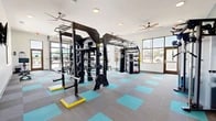 Located at Covington Heights Circle in Winston-Salem, NC, this fitness center offers a comprehensive and thoughtfully outfitted workout environment spread across a single open floor. The space is defined by its modern, clean aesthetic, featuring bright white walls accented by a teal-painted feature wall, and a distinctive two-tone carpet tile floor in gray and turquoise that adds a dynamic visual energy to the room. Multiple ceiling fans and recessed lighting ensure the space remains comfortable and well-lit throughout. Large windows and glass garage-style doors line the perimeter, inviting abundant natural light and providing views of the surrounding outdoor environment. The equipment selection is extensive and designed to support a wide variety of training styles. A central multi-functional strength rig serves as the focal point, accommodating barbell work, suspension training, and pull-up movements. Nearby, a dedicated free weight area features a full rack of dumbbells and kettlebells alongside adjustable benches. TKO-branded punching bags and a functional trainer cable machine support combat and cross-training workouts. The cardio zone houses multiple treadmills and rowing machines. Wall-mounted flat-screen televisions add an entertainment element, while the overall layout provides ample open floor space for functional and stretching exercises.