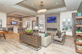 Located at 4590 N Campbell Rd in Royal Oak, Michigan, this single-floor clubhouse showcases a thoughtfully designed contemporary interior that blends comfort and sophistication. The entry foyer sets a striking first impression with large-format light wood tile flooring laid in a herringbone pattern and a bold, angular decorative screen partition that visually separates spaces while maintaining an open, airy feel. Recessed lighting and pendant fixtures illuminate the space throughout.

The main lounge area is a standout, featuring tray ceilings with elegant chandelier lighting, a wall-mounted linear gas fireplace flanked by textured stone tile, and a large flat-screen TV. Plush neutral seating, teal accent pillows, layered area rugs, and open shelving with curated décor create a warm yet modern atmosphere. A secondary seating nook provides additional gathering space.

A dedicated kitchenette area offers dark cabinetry, white countertops, stainless steel appliances, and a commercial-grade coffee station, all set against a rustic reclaimed wood accent wall with a mounted display screen. A game area with a foosball table adds a recreational element.

Natural light filters through multiple windows and glass entry doors, enhancing the spacious, welcoming ambiance of this well-appointed community space.